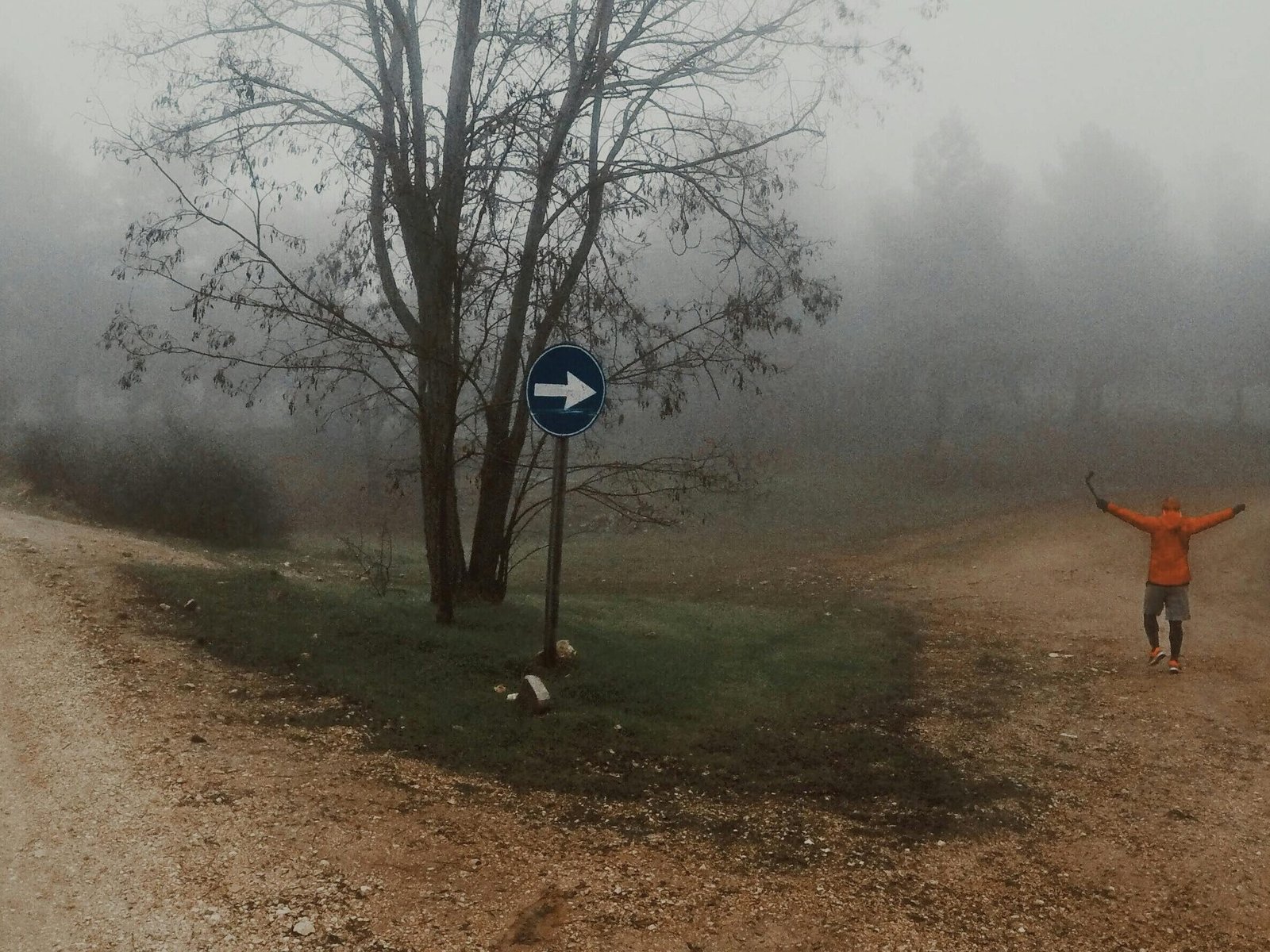 A person in an orange jacket explores misty dirt roads in a foggy forest setting.